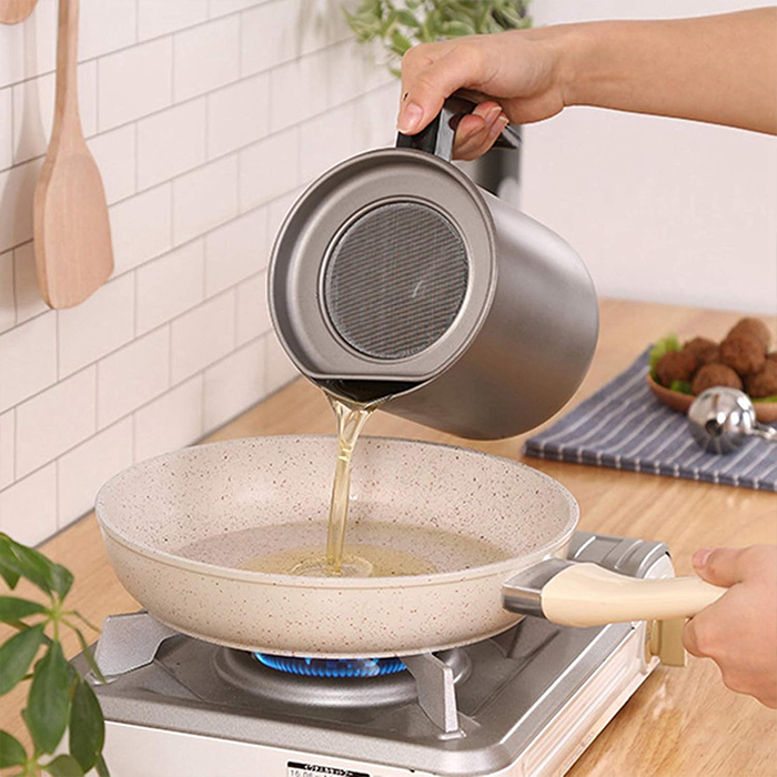 A person is pouring filtered oil from a stainless steel container into a frying pan.