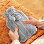 Soft grey hot water bottle with plush cover and pom-pom ties, ideal for soothing aches, warming hands and feet, or use as a cold compress.