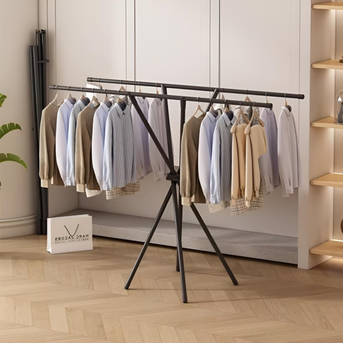 X-Shaped Folding Clothes Drying Rack made of stainless steel, with clothes hanging on it, placed on a wooden floor against a white wall.